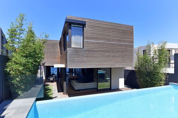 A modern multi-level residential house viewed from the pool, featuring a cantilevered upper volume clad in horizontal dark timber boards, expansive ground-floor glazing, and a clean rectilinear form. The bright blue swimming pool dominates the foreground, with landscaped planting and neighbouring contemporary buildings visible around the site.