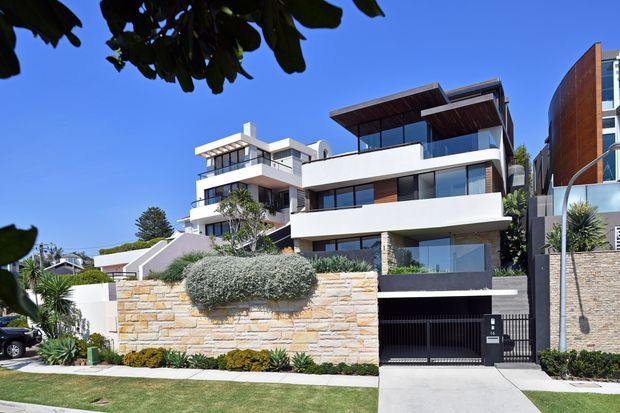 Street-facing view of a contemporary multi-storey residential building with crisp white rendered volumes, extensive glazing, dark metal framing, and projecting rooftop terraces with timber-lined soffits. A layered natural-stone boundary wall, landscaped planting, driveway, and garage entry occupy the foreground. The bright blue sky and mature foliage frame the modern facade.