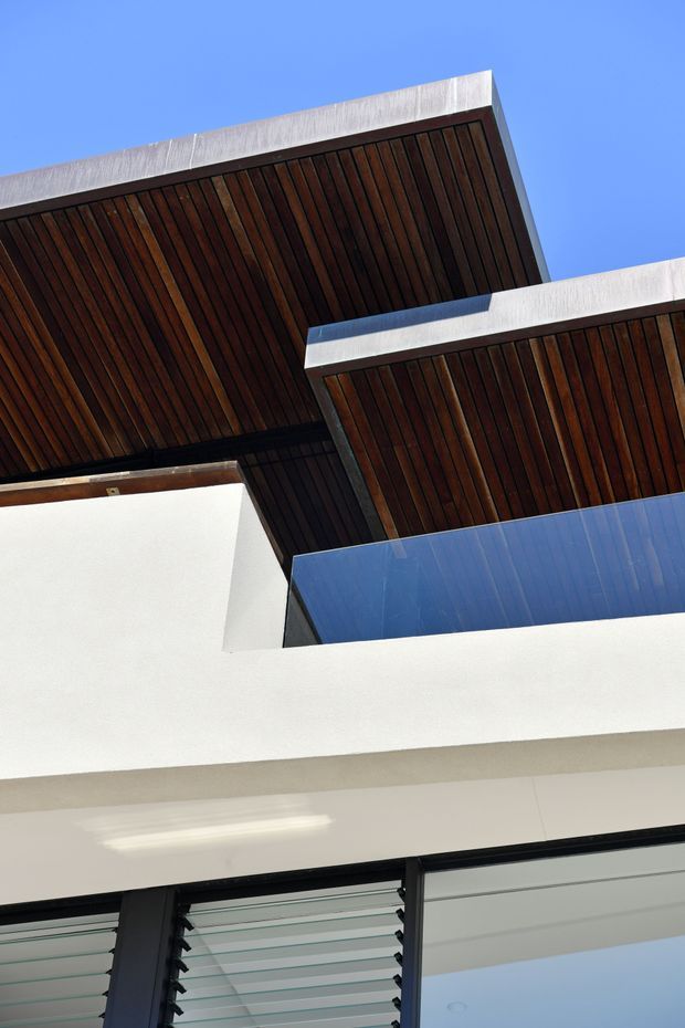 Upward-looking architectural detail of a contemporary multi-level building, featuring projecting flat roof planes with dark timber-lined soffits, crisp white rendered walls, black-framed glazing, and horizontal louvres against a clear blue sky.