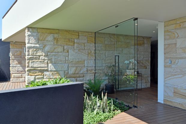 Modern residential entrance viewed from an exterior deck, featuring extensive horizontally coursed natural stone walling, frameless glass entry doors, a white projecting flat roof, dark rendered planter walls, timber decking, and integrated planting. The composition emphasises a sheltered, contemporary indoor-outdoor threshold.