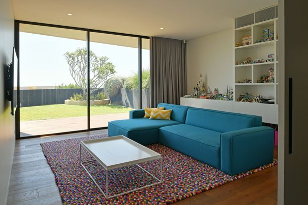 Bright contemporary living room with a large teal-blue sectional sofa, low rectangular coffee table, and colourful patterned rug. Floor-to-ceiling black-framed sliding glass doors open to a landscaped courtyard, while built-in white shelving and cabinetry line the right wall. Neutral walls, recessed ceiling lighting, timber flooring, and full-height curtains create a clean, family-oriented interior.