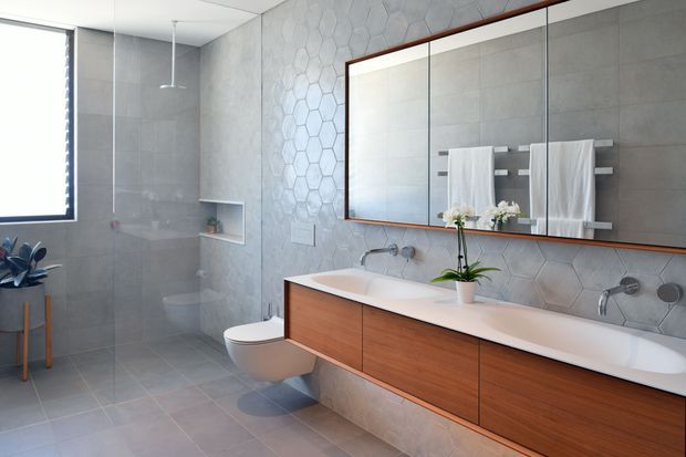 Contemporary bathroom with a long floating timber vanity, dual integrated basins, wall-mounted chrome taps, a large framed mirrored cabinet, and twin towel rails. The room features pale grey tiled walls and floor, a hexagonal feature-tile wall, a frameless glass shower enclosure, wall-hung toilet, and a small window providing natural light.