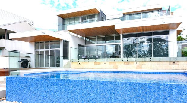 Exterior view of a large contemporary multi-level residence with extensive glazing, crisp white rendered walls, projecting flat roof planes and warm timber soffits. A glass-balustraded terrace overlooks a prominent blue mosaic-tiled swimming pool in the foreground.