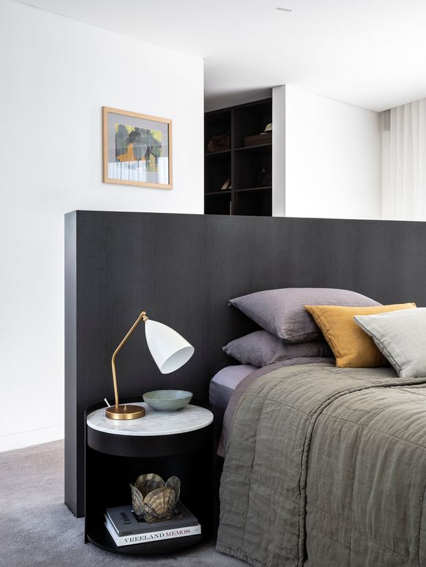 A contemporary bedroom with a low dark built-in partition and integrated open shelving behind the bed. The room features a layered grey bedspread and pillows with mustard and pale grey accents, a round black bedside table with a light stone top, and a brass-and-white table lamp. White walls, pale carpet, sheer curtains, and a framed artwork create a clean, refined interior.