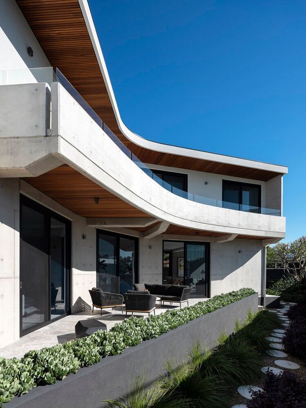 Modern two-storey residential exterior with sweeping curved balconies, exposed concrete walls, dark-framed glazing, and warm timber-lined soffits. The building opens onto a landscaped terrace and narrow garden path under a clear blue sky.