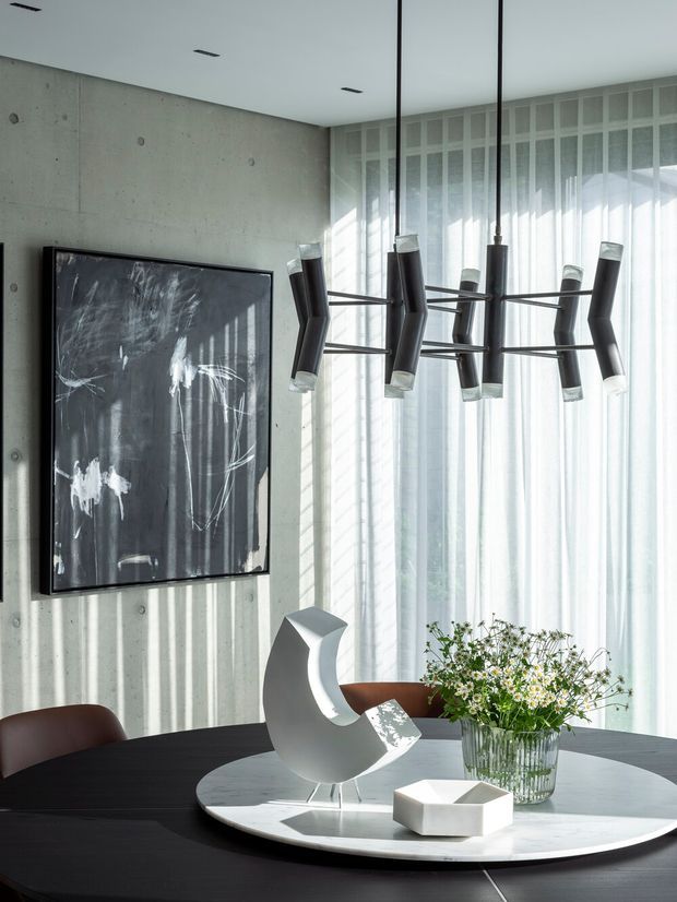 A sophisticated contemporary dining area with a dark round dining table, sculptural white centrepiece, flower arrangement, and curved brown dining chairs. The room features a raw concrete feature wall, expansive sheer-covered glazing, and a striking linear black pendant light with multiple tubular shades. A large monochrome artwork adds an artistic focal point.