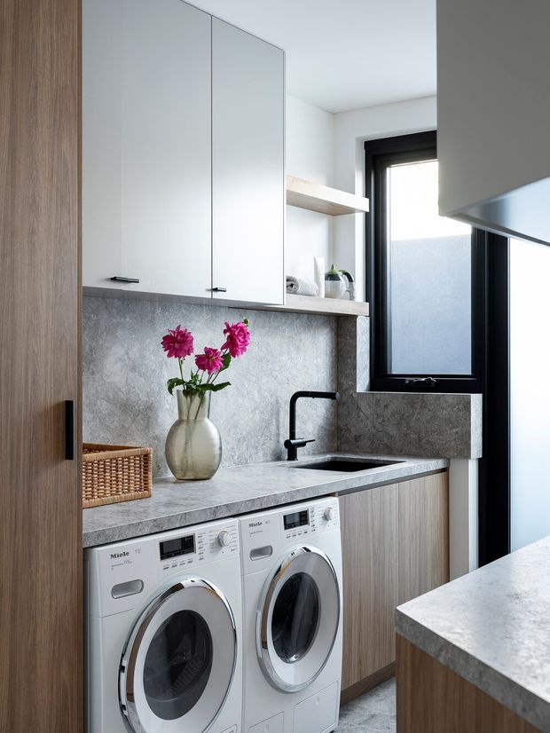 A compact, contemporary laundry room with stacked side-by-side front-loading Miele washer and dryer beneath a grey stone-look benchtop. The room features light timber cabinetry, white overhead cupboards, a black-framed window and black tapware, a grey textured splashback, open shelving, and a deep undermount sink. A light stone benchtop from adjacent cabinetry is visible in the foreground.