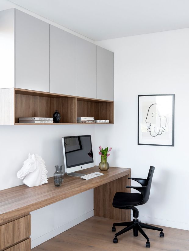 Minimal built-in home office with a wall-mounted timber desk, integrated wood shelving, pale grey overhead cabinetry, white walls, and a black task chair. An iMac, books, decorative objects, and framed line-art add functional and restrained styling.