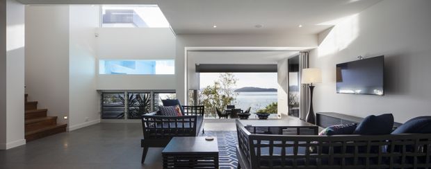 Bright contemporary open-plan living room with a double-height ceiling, expansive glazing, and a wide view toward the water. The room contains dark timber-framed sofas, a low coffee table, a wall-mounted television, a dining area near the glazing, and a timber staircase at left. White walls and polished grey flooring create a clean, airy interior.