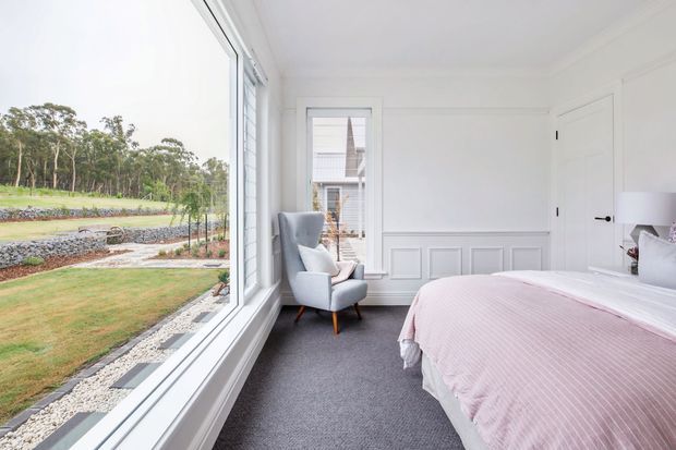 Bright, refined bedroom with a large corner window overlooking landscaped grounds. The room features white walls, decorative lower wall panelling, a grey carpet, a pale upholstered armchair, a partially visible bed with a soft pink cover, and a white paneled door. The interior has a classic, airy character with abundant natural light.