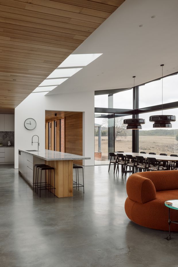 Open-plan contemporary interior combining a kitchen, dining area and living space. A large stone-topped island with timber-clad sides and black stools anchors the kitchen, while full-height glazing opens to a rural landscape. The room features a polished concrete floor, white walls, extensive timber ceiling panelling, skylights, dark window frames, sculptural pendant lights, a dining table, and a curved rust-orange sofa.