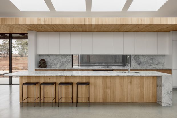 Contemporary open-plan kitchen dominated by a large timber island with four slim bar stools, pale stone benchtops, and a dramatic grey-veined stone splashback. Full-height white cabinetry lines the rear wall, while warm timber ceiling lining and rectangular skylights add texture and natural light. Large glazing opens to a sparse rural landscape.