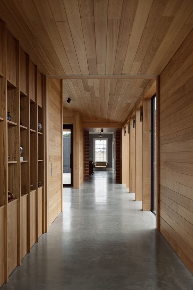 A long, refined residential hallway lined with warm timber wall and ceiling panelling. Built-in timber shelving and cabinetry run along the left wall, while timber-lined openings and dark-framed glazing define the right side. The polished grey concrete floor leads toward a brick-framed sitting area with a large window at the far end. The space has a calm, minimalist material palette and strong linear perspective.