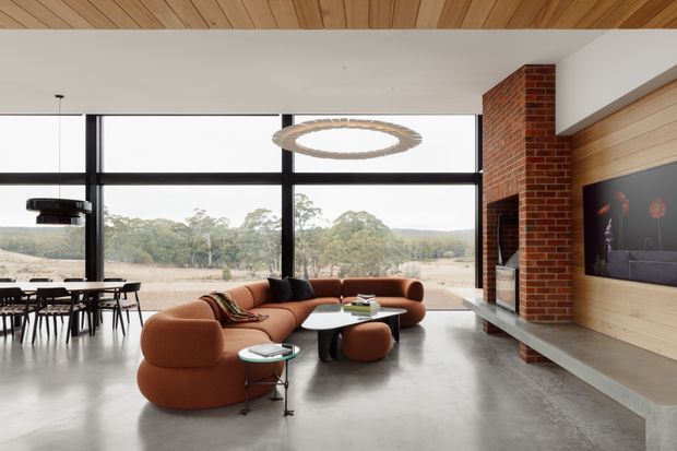 A spacious contemporary living room with floor-to-ceiling black-framed glazing overlooking a rural landscape. The room features a curved rust-brown sectional sofa, round coffee tables, sculptural pendant lighting, polished concrete flooring, a timber-lined ceiling and feature wall, and an exposed red-brick fireplace with a long concrete hearth. A dining area is visible to the left.