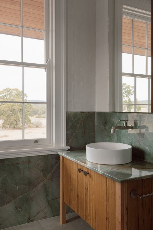 A refined bathroom vanity area with a white vessel basin on a timber vanity, green veined stone benchtop and backsplash, white painted wall panelling, and traditional multi-pane windows with timber roller blinds. A wall-mounted brushed metal tap and mirrored reflection are visible, with a muted natural landscape beyond the windows.