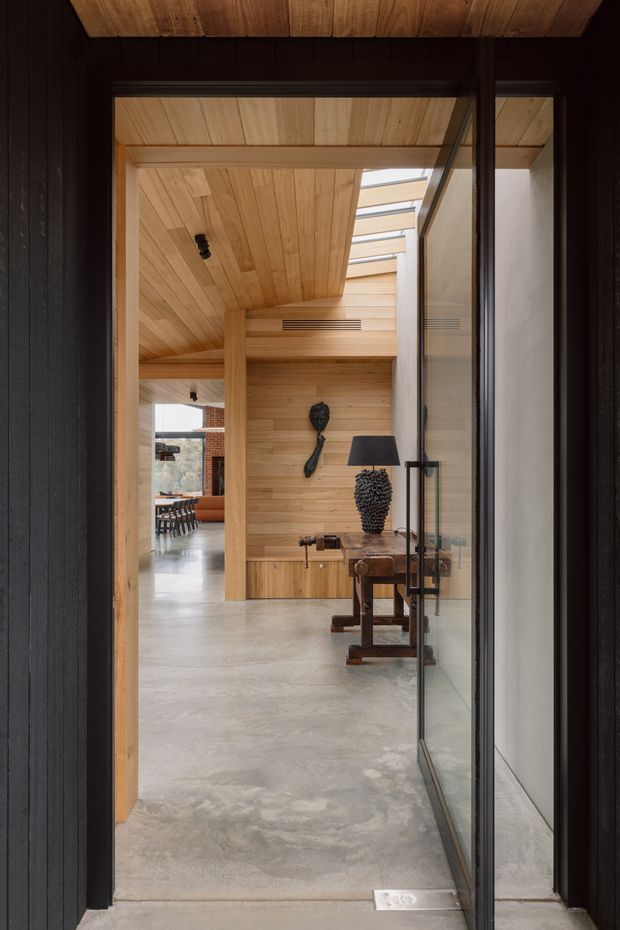 View through a dark-framed glazed entry door into a warm, timber-lined interior. The space features extensive light timber wall and ceiling panelling, a polished grey concrete floor, a skylit corridor or entry hall, and a sculptural console table with dark decorative objects. A dining or living area is visible beyond.