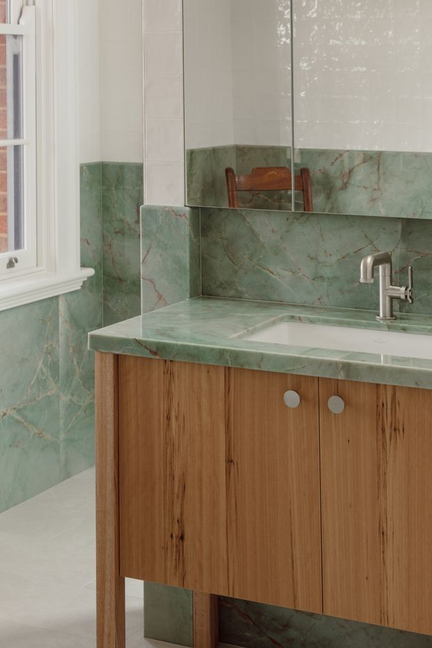 A compact bathroom vanity with a warm timber cabinet, integrated white basin, brushed metal tapware, and green veined stone used across the benchtop and wall surfaces. A large mirrored cabinet reflects the room, while a white-framed window provides natural light. The palette is soft green, warm wood, white, and muted metal.