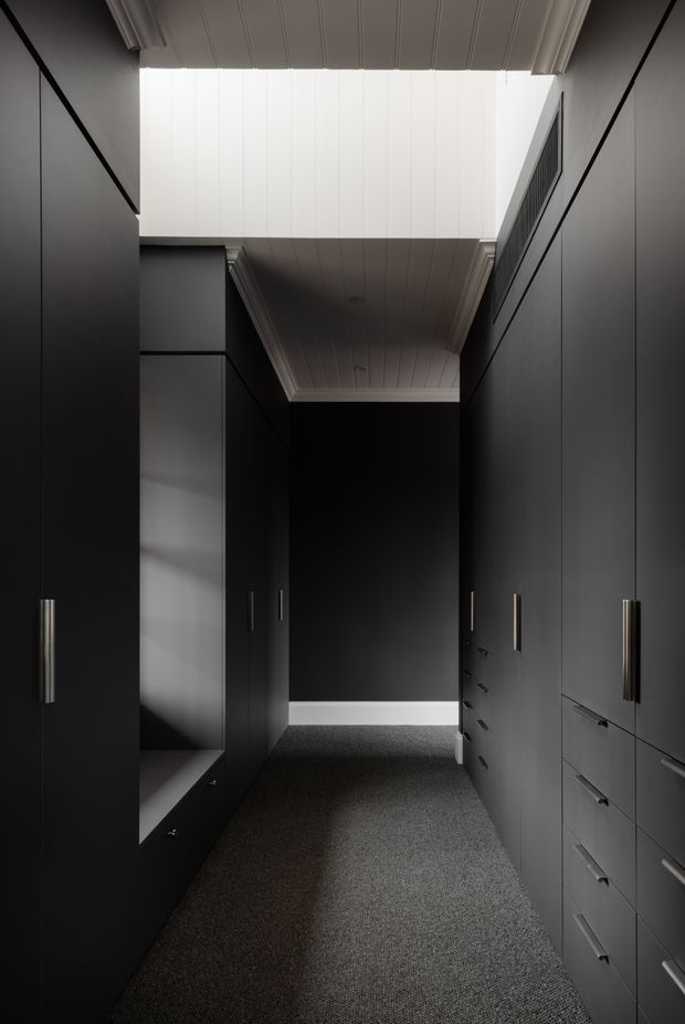 A narrow, dark walk-in wardrobe lined with full-height matte black cabinetry and long slim metal handles. The space has grey carpet flooring, integrated drawers and shelving, and a white vertically panelled ceiling with a bright clerestory-like opening that brings in natural light. The overall composition is highly minimalist and monochromatic with strong linear detailing.
