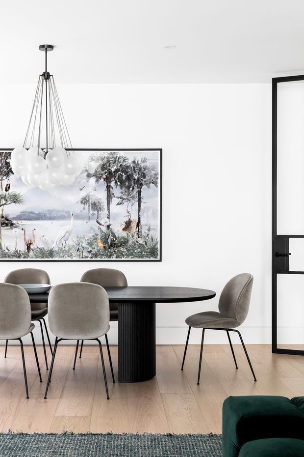 A bright, refined dining area with a round black dining table and sculptural central pedestal, surrounded by upholstered taupe dining chairs. Pale timber flooring, white walls, a large framed botanical artwork, a sculptural pendant light, and a black-framed glazed door create a contemporary monochromatic interior. A dark green upholstered sofa is partially visible in the foreground.