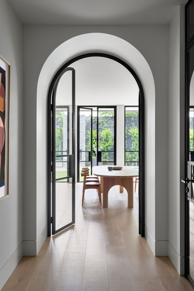 A bright, refined interior view through a large rounded archway with black metal-framed glazed doors. Beyond the opening is a dining area with a round timber table, curved timber legs, and matching wooden chairs, surrounded by large black-framed windows and greenery outside. Pale timber flooring, white walls, and a sculptural arched doorway create a clean contemporary character.