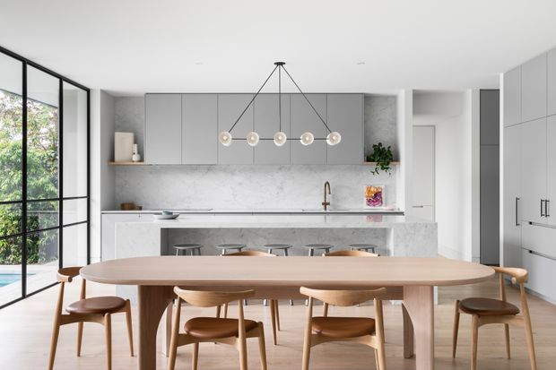 Bright, refined open-plan kitchen and dining area with a long light timber dining table and matching wooden chairs in the foreground. The kitchen features matte grey floor-to-ceiling cabinetry, a pale stone island and benchtop, a light veined stone splashback, integrated appliances, and a row of pendant lights. Black-framed full-height glazing opens toward greenery, while pale timber flooring and white walls create an airy contemporary setting.