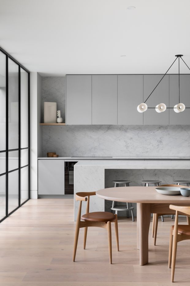 A refined open-plan kitchen and dining area with a minimalist grey and white palette. The kitchen features full-height matte grey cabinetry, a veined stone backsplash and island, integrated appliances, and a slim linear pendant with circular shades. A light timber dining table and matching timber chairs occupy the foreground, while black-framed floor-to-ceiling glazing lines the left side. Pale timber flooring and restrained detailing create a calm, contemporary interior.