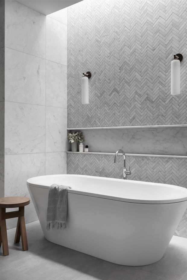 A refined contemporary bathroom featuring a freestanding white bathtub, chrome wall-mounted faucet, large pale marble-look wall tiles, and a contrasting herringbone mosaic tile feature wall. Slim recessed shelves, cylindrical wall sconces, and a small timber stool complete the calm monochromatic scheme.