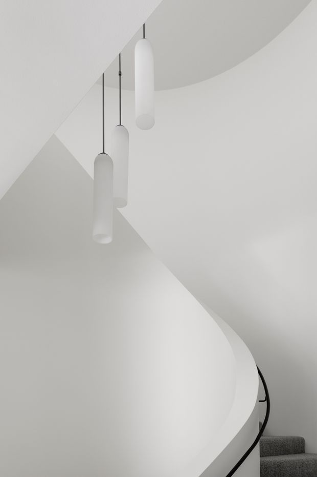 Minimalist architectural interior showing a sculptural curved staircase with smooth white plaster walls and balustrade, dark handrail, grey carpeted treads, and three suspended cylindrical pendant lights. The composition emphasises flowing geometry, a monochromatic palette, and soft diffused light.