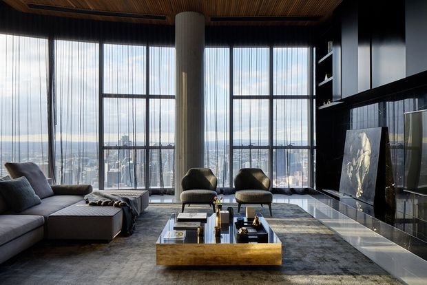 A sophisticated high-rise living room with floor-to-ceiling windows overlooking a dense city skyline. The room features a large sectional sofa, sculptural lounge chairs, a substantial coffee table, dark built-in cabinetry and media joinery, a dramatic artwork panel, a cylindrical column, and a richly textured timber ceiling with recessed linear detailing. The palette is dark and refined with warm wood accents and abundant daylight.