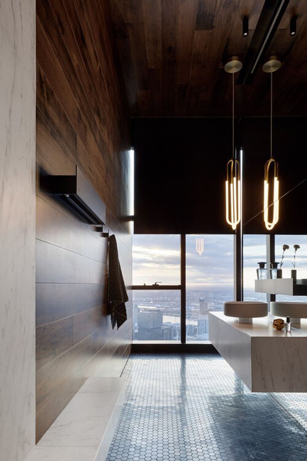 A sophisticated contemporary bathroom with a floating double vanity, large mirror, wall-mounted television, freestanding vessel basins, and sculptural pendant lighting. The room combines dark timber wall and ceiling lining with pale marble-look surfaces and a small-format blue-grey mosaic tile floor. Full-height glazing provides expansive city views, while integrated storage and minimal detailing create a luxurious high-rise feel.