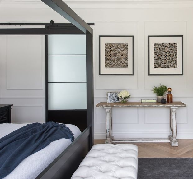 A refined bedroom interior featuring a black four-poster or canopy bed with a dark blue throw, white paneled walls, and a black-framed frosted-glass sliding door. A weathered timber console table with decorative objects sits beneath two framed geometric artworks, while a tufted white bench and dark rug complete the room. The space combines classic wall detailing with contemporary dark accents and warm timber flooring.