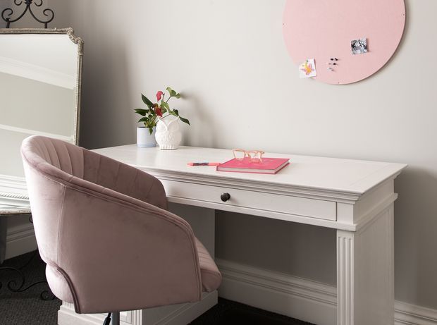 A softly styled home office or study corner featuring a white classic writing desk with fluted leg detailing and a drawer, paired with a blush pink upholstered chair. A large ornate mirror, decorative vase with greenery, and pink circular pinboard add feminine decorative accents. The neutral walls and white trim create a refined, light atmosphere.