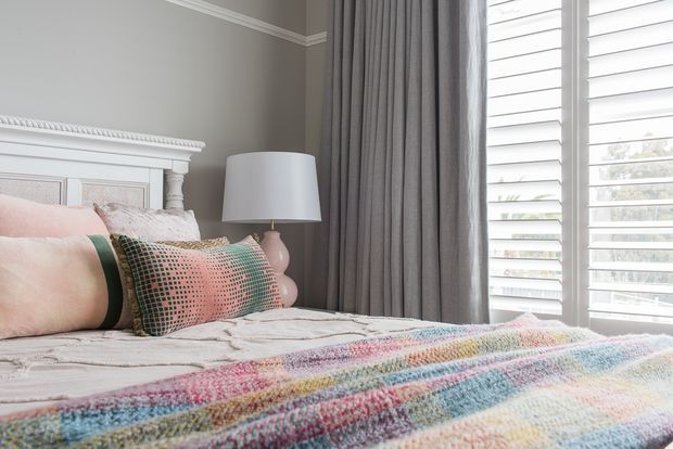 A softly lit, traditionally styled bedroom with a white decorative paneled bedhead, layered pale bedding, patterned cushions, and a multicoloured textured throw. Grey curtains frame a large window with white plantation shutters, while a simple bedside lamp adds warmth.