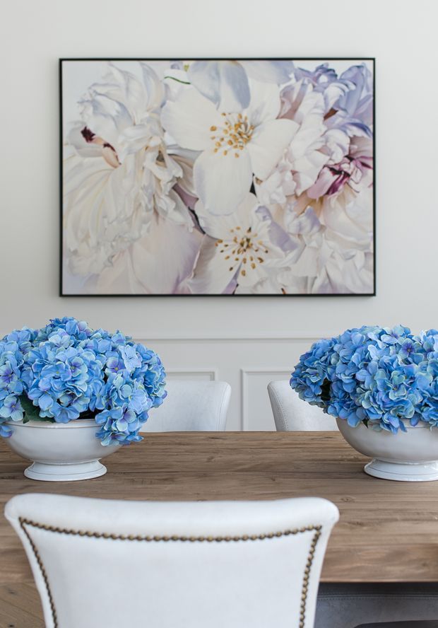 A refined dining room featuring a large wooden dining table surrounded by upholstered white chairs with dark nailhead trim. Two large white pedestal bowls filled with blue hydrangeas form a symmetrical centerpiece, while a framed pastel floral artwork hangs above white decorative wall panelling.