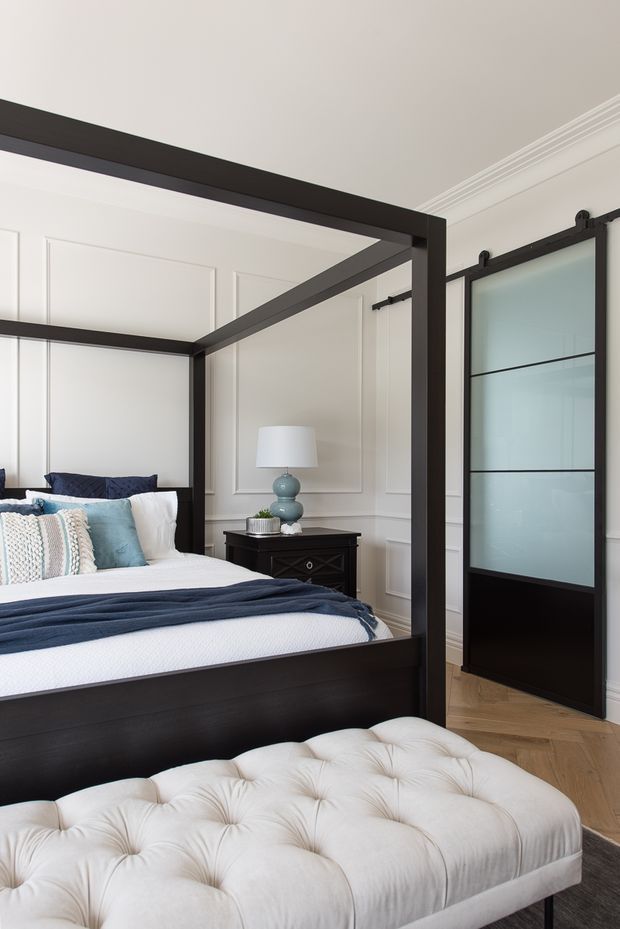 A refined bedroom featuring a dark timber four-poster bed, white bedding with navy and pale blue accents, a tufted cream upholstered bench, decorative white wall panelling, and a black-framed frosted-glass sliding barn door. Light timber flooring and traditional ceiling cornices add architectural detail.
