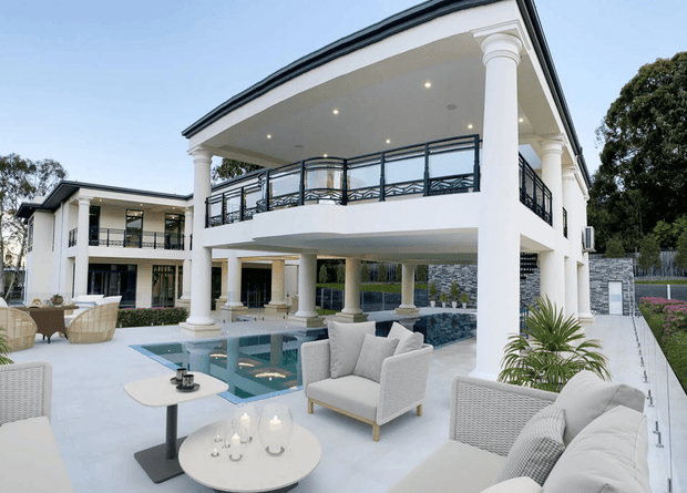 A luxurious two-storey white rendered residence with a large covered outdoor entertaining area, classical columns, dark metal balustrades, and expansive poolside terraces. The foreground contains pale outdoor lounge furniture and coffee tables, while the architecture features an open upper balcony and a substantial swimming pool integrated into the terrace.