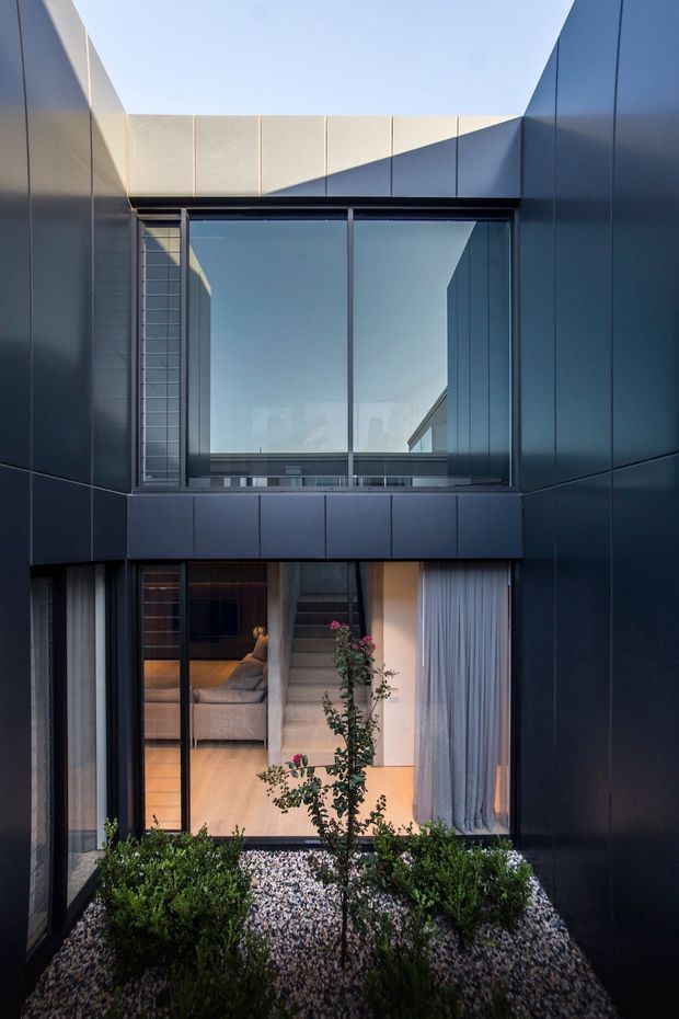A narrow, two-storey modern courtyard viewed from outdoors, enclosed by dark blue-grey metal-clad walls. Large floor-to-ceiling glazed openings reflect the sky and surrounding structure, while the lower opening reveals a warm living room with a sofa, timber flooring and full-height curtains. A minimalist gravel courtyard with clipped shrubs and a small ornamental tree occupies the foreground. Strong geometric lines, a restrained palette and dramatic evening-like light create a refined contemporary architectural composition.