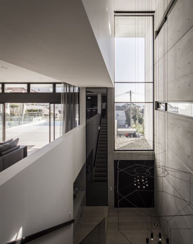 A dramatic interior view of a contemporary multi-level stair hall or landing. A narrow dark staircase descends beside a tall void with floor-to-ceiling glazing, while a raw concrete feature wall with visible formwork joints and a delicate geometric pendant light dominates the right side. The adjacent open-plan living area and outdoor pool or courtyard are visible through glazing on the left. The space has strong linear geometry, abundant natural light, and a restrained monochromatic palette.