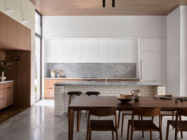 A refined open-plan kitchen and dining area with a long timber dining table and mid-century-style wooden chairs in the foreground. The kitchen features a white tiled island, white flat-panel cabinetry, a grey marble-look splashback and benchtops, timber cabinetry, and a timber-lined ceiling. A dark timber built-in wall with shelving and cabinetry sits beside tall windows, while polished grey flooring and contemporary linear lighting complete the space.