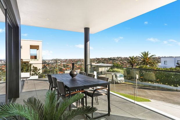 A contemporary covered outdoor terrace with a dark dining table and chairs, glass balustrade, tiled floor, and a substantial structural column. The open view overlooks a sunny suburban landscape with palm trees and neighbouring homes, while large glazed openings connect the terrace to the interior.