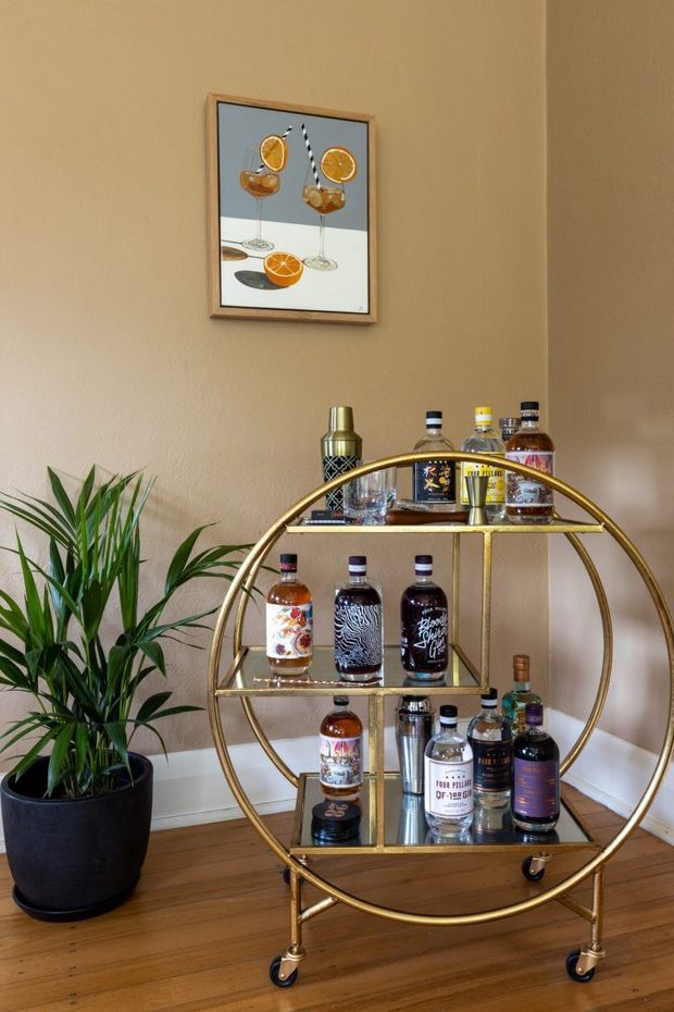 A styled home bar corner featuring a circular brass-gold bar cart with multiple shelves, bottles, cocktail-making accessories, and caster wheels. The cart sits against a warm beige wall above timber flooring, with a tall potted plant beside it and framed cocktail artwork above.