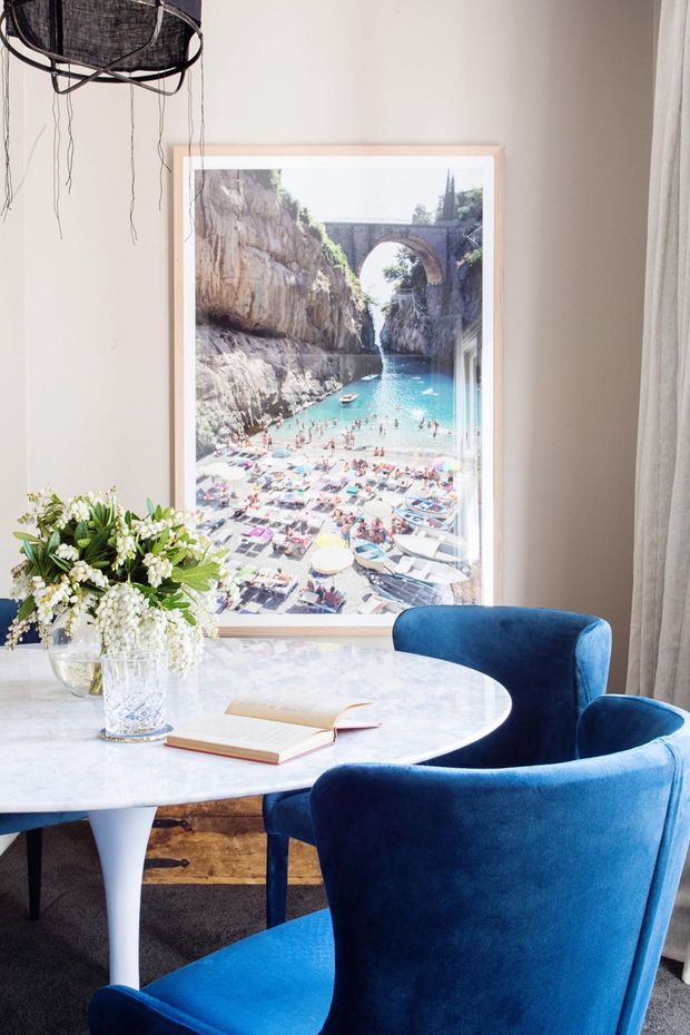 A bright, styled dining area featuring a round white marble table, two plush deep-blue upholstered armchairs, and a large framed coastal travel photograph. A floral arrangement, open book, textured pendant light, curtains, and dark carpet complete the refined interior.