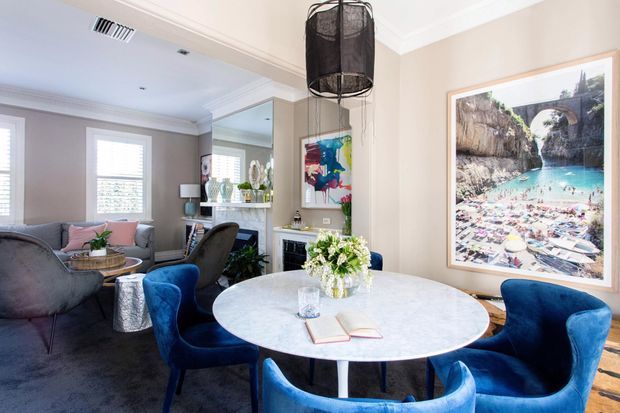 A bright, refined open-plan dining and living area. The foreground features a round white stone-look dining table with vivid blue upholstered dining chairs, while a grey lounge seating area, wall mirror, artwork, decorative lighting, and built-in shelving occupy the background. Neutral walls, white trim, dark carpet, and timber flooring create a polished contemporary interior.