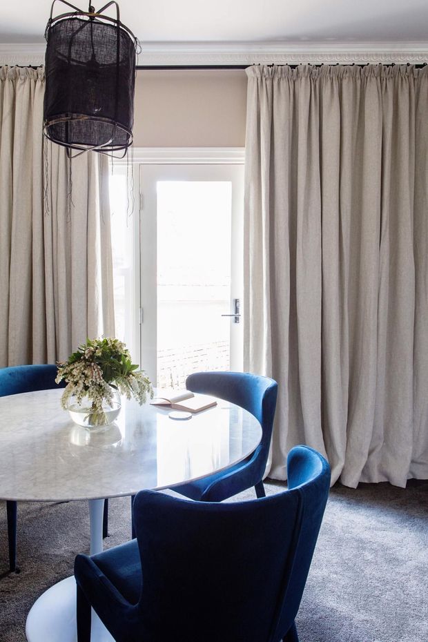 Elegant dining room with a round pale stone or marble-topped table, deep blue upholstered dining chairs, full-height neutral curtains, a white glazed door, decorative ceiling cornice, and an oversized sculptural dark pendant light. The room has a refined, softly lit atmosphere with a restrained neutral-and-blue palette.