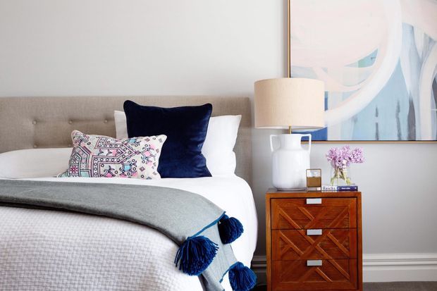 A softly styled bedroom with a tufted upholstered headboard, white bedding, a grey throw with navy tassels, patterned and navy accent pillows, and a warm timber bedside chest. A ceramic lamp, flowers, and abstract artwork add decorative detail against pale walls with traditional wall trim.