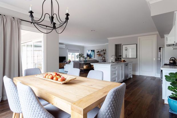 Bright open-plan dining and kitchen interior with a large light timber dining table, upholstered dining chairs, white cabinetry, dark timber flooring, and a black decorative chandelier. The kitchen includes a central island, extensive built-in storage, and pale benchtops, with a living area visible beyond.