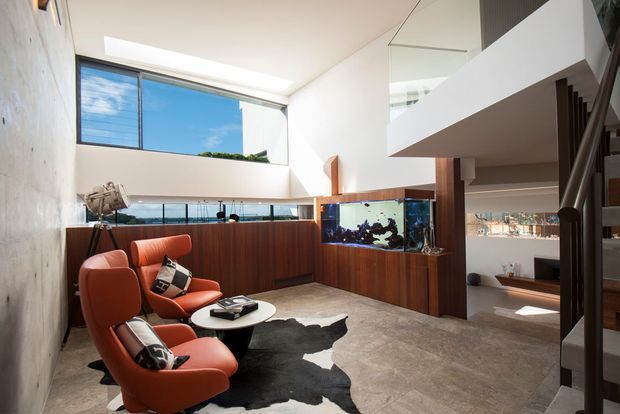 Bright double-height contemporary living space with a dramatic concrete feature wall, expansive glazing, and an upper-level void. The room includes sculptural orange lounge chairs, a low round coffee table, a large aquarium integrated into dark timber cabinetry, polished concrete flooring, and a staircase at right.