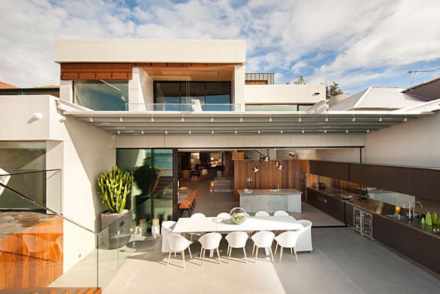 A bright contemporary multi-level home with a large covered outdoor entertaining area. The scene features a long dining table with white chairs, an outdoor kitchen with dark cabinetry and a stone island, extensive glazing, timber wall cladding, and a rooftop terrace or balcony above. Clean geometric lines and pale finishes create a spacious indoor-outdoor living environment.
