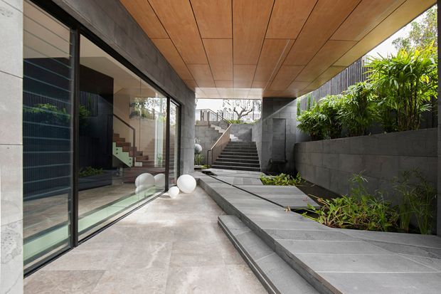 A modern internal courtyard or covered outdoor passage with large black-framed glazed doors on the left, a warm timber-lined soffit overhead, and dark grey masonry walls and stepped planters on the right. Pale stone paving leads toward a central staircase, with lush planting providing greenery along the raised garden bed. The glazing reveals an interior staircase and furnishings beyond.
