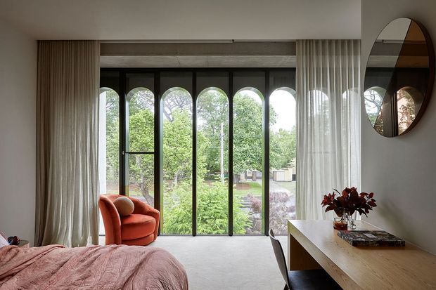 A refined bedroom with floor-to-ceiling arched glazing overlooking a leafy garden. The room includes a low-profile bed with blush bedding, a burnt-orange lounge chair, sheer full-height curtains, a pale wood desk or console, and a large round wall mirror. The palette is soft and warm, with strong natural light and a contemporary, understated interior.
